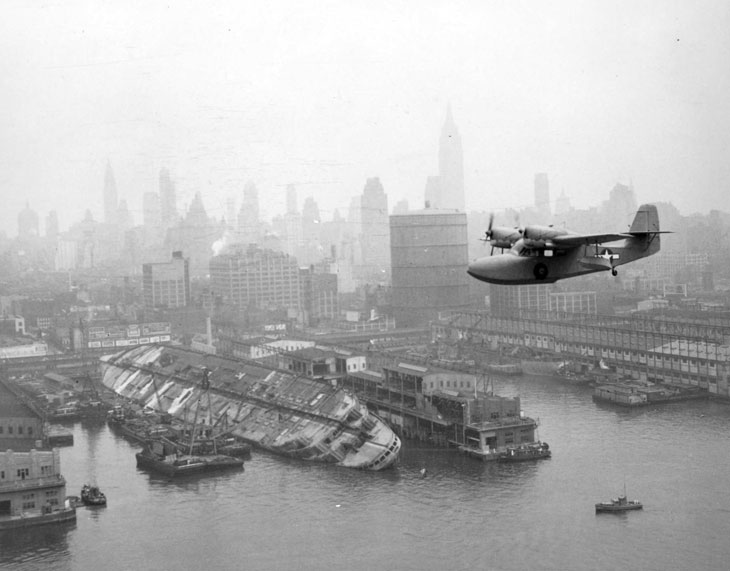 A U.S. Coast Guard Grumman J4F Widgeon flies over the wreckage of the USS Lafayette (AP-53) at Pier 88, New York harbor, on 12 August 1943