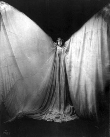 Loïe Fuller standing, facing front; wearing large gown, with arms raised in shape of a butterfly.