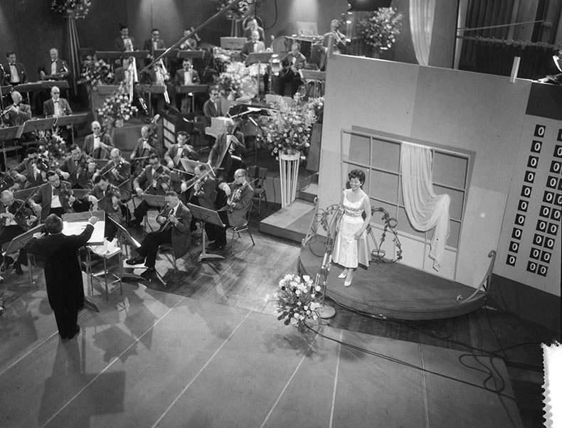 Black and white photograph of a female singer in a white dress performing on stage with a live band 