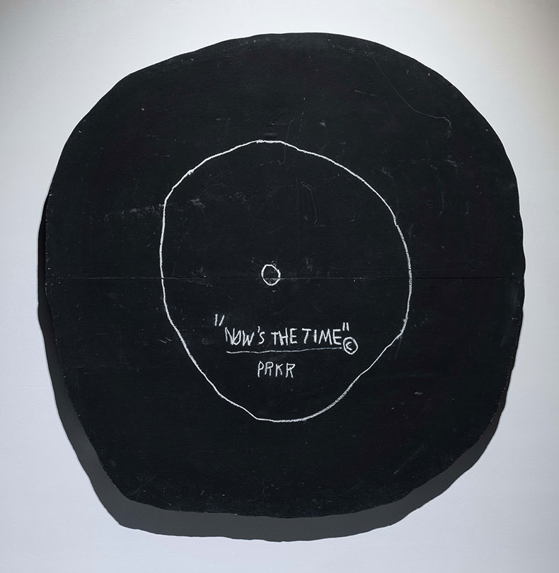 a black circular painting with words scratched in the centre reading Now’s the Time